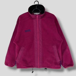 May include: A pink fleece jacket with a zip fastening and a drawstring at the bottom. The jacket has a small logo on the left chest.