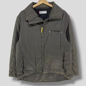 Puede incluir: Chaqueta acolchada verde oliva con cuello alto y cremallera completa. La chaqueta tiene un corte cuadrado, dos bolsillos laterales y una pequeña etiqueta con el logotipo cerca del cuello. La chaqueta está colgada de una percha de madera.