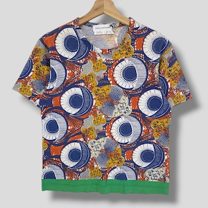 K&ouml;nnte beinhalten: Kurz&auml;rmeliges T-Shirt mit einem auff&auml;lligen Muster aus blauen, orangefarbenen, wei&szlig;en und gelben Kreisen und floralen Designs. Das Shirt hat einen gr&uuml;nen Saum und h&auml;ngt an einem Holzb&uuml;gel.