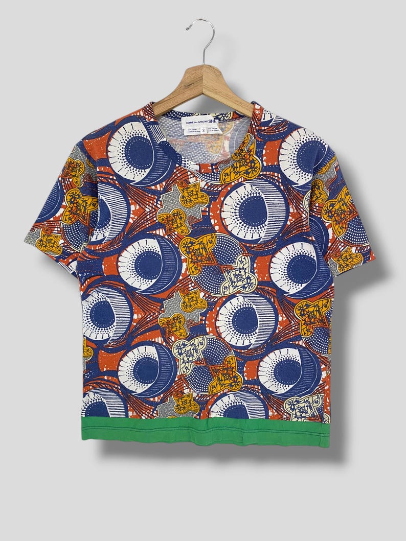K&ouml;nnte beinhalten: Kurz&auml;rmeliges T-Shirt mit einem auff&auml;lligen Muster aus blauen, orangefarbenen, wei&szlig;en und gelben Kreisen und floralen Designs. Das Shirt hat einen gr&uuml;nen Saum und h&auml;ngt an einem Holzb&uuml;gel.