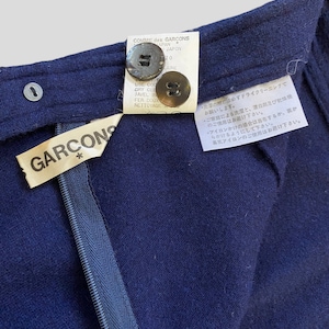 Puede incluir: Primer plano de una prenda azul marino con cremallera y dos botones. La prenda tiene una etiqueta que dice "GARCONS" y otra etiqueta con texto en japon&eacute;s y franc&eacute;s. Los botones son marrones y la cremallera es azul.