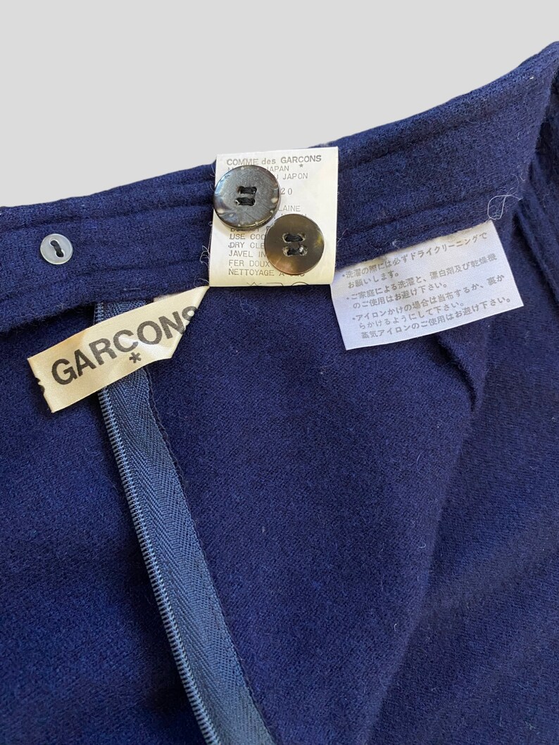 Puede incluir: Primer plano de una prenda azul marino con cremallera y dos botones. La prenda tiene una etiqueta que dice "GARCONS" y otra etiqueta con texto en japon&eacute;s y franc&eacute;s. Los botones son marrones y la cremallera es azul.