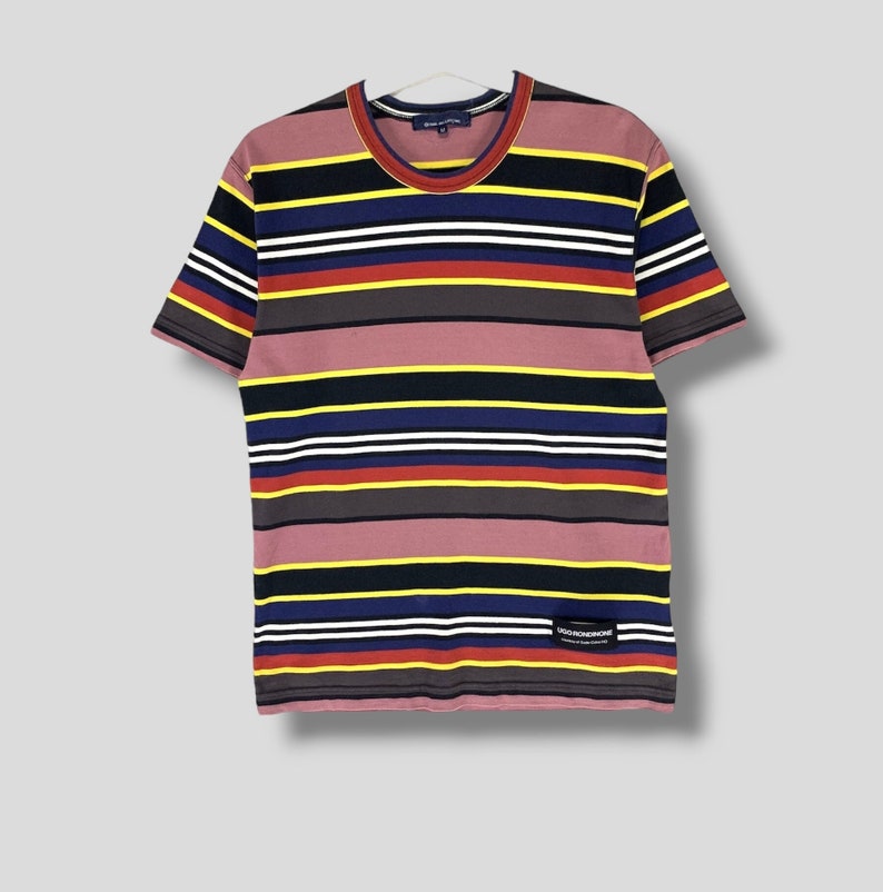 May include: A short-sleeved t-shirt with horizontal stripes in various colors, including pink, black, yellow, blue, red, and white. The shirt has a crew neck and a small label near the bottom. The shirt is hanging on a white hanger.