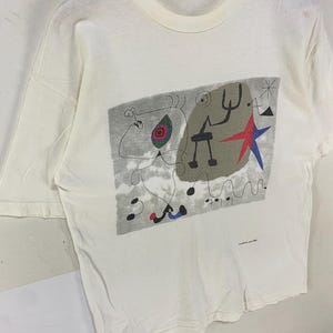 Puede incluir: Camiseta de color crema con un dise&ntilde;o abstracto colorido. El gr&aacute;fico incluye una estrella roja, formas negras y un fondo gris. La camiseta est&aacute; colgada en una percha de madera.