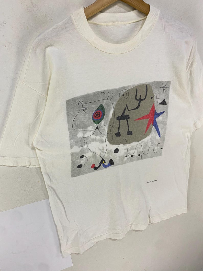 Puede incluir: Camiseta de color crema con un dise&ntilde;o abstracto colorido. El gr&aacute;fico incluye una estrella roja, formas negras y un fondo gris. La camiseta est&aacute; colgada en una percha de madera.