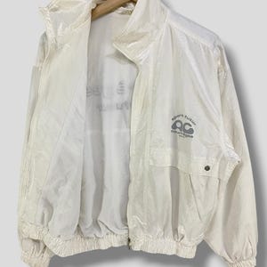 K&ouml;nnte beinhalten: Cremefarbene Vintage-Windjacke mit hohem Kragen und durchgehendem Rei&szlig;verschluss. Die Jacke hat eine kleine Tasche und den Text "sport futur courreges" in Grau.
