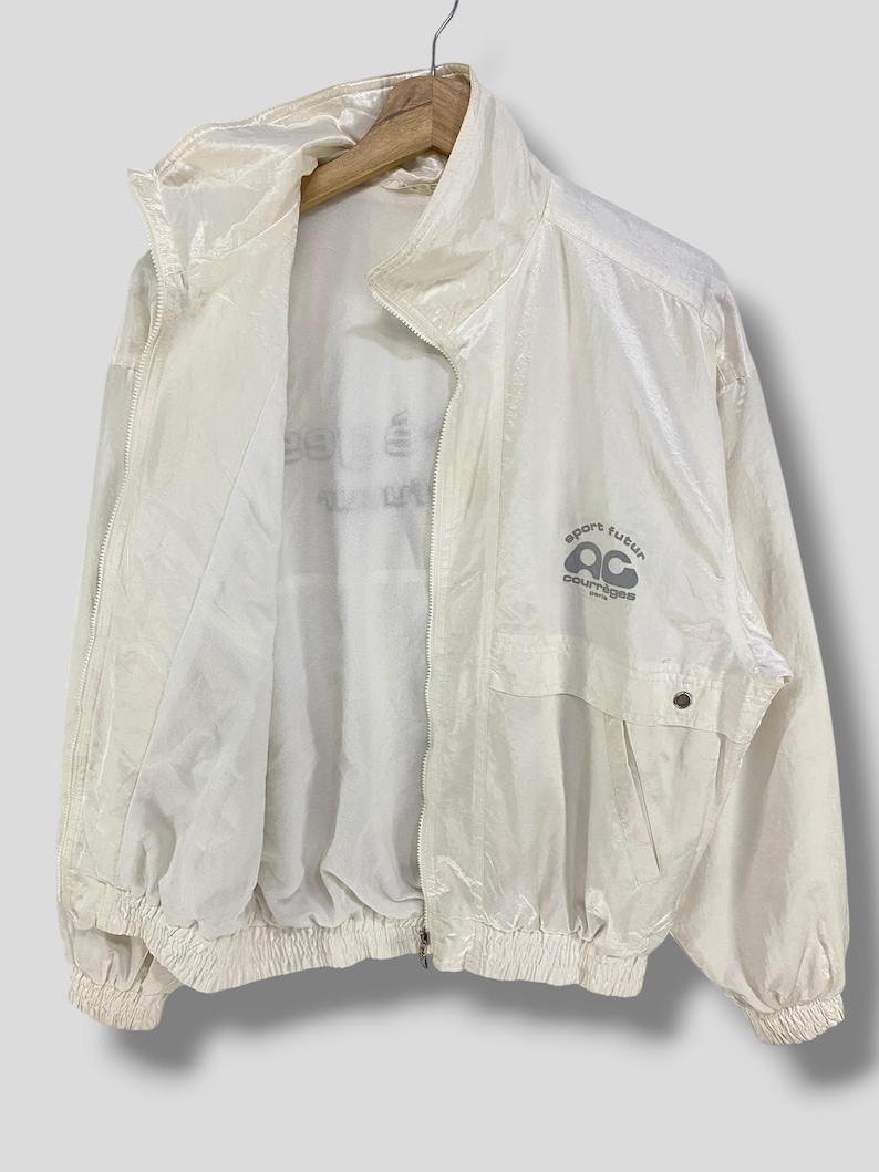 K&ouml;nnte beinhalten: Cremefarbene Vintage-Windjacke mit hohem Kragen und durchgehendem Rei&szlig;verschluss. Die Jacke hat eine kleine Tasche und den Text "sport futur courreges" in Grau.