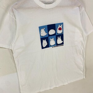May include: White t-shirt featuring a graphic of six cartoon characters in a blue and white square. The text "MY NEIGHBOR TOTORO" is printed below the graphic. The shirt is hanging on a wooden hanger.