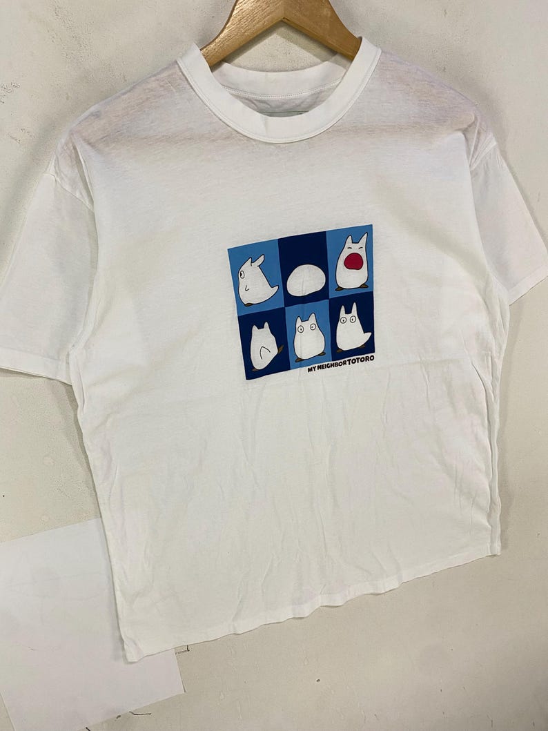 May include: White t-shirt featuring a graphic of six cartoon characters in a blue and white square. The text "MY NEIGHBOR TOTORO" is printed below the graphic. The shirt is hanging on a wooden hanger.