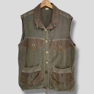 May include: A sleeveless olive green vest with a brown collar and accents. The vest has two front pockets, decorative stitching, and a drawstring at the bottom. It is buttoned up the front and hangs from a wooden hanger.