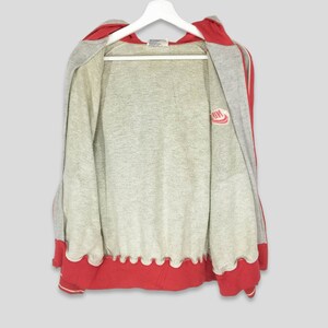 K&ouml;nnte beinhalten: Ein grauer Kapuzenpullover mit Rei&szlig;verschluss und roten Akzenten, der an einem wei&szlig;en Kleiderb&uuml;gel h&auml;ngt. Die Kapuze und der Saum sind rot, mit einem wei&szlig;en Streifen. Das Nike-Logo ist auf der Brust zu sehen. Die Jacke ist offen.