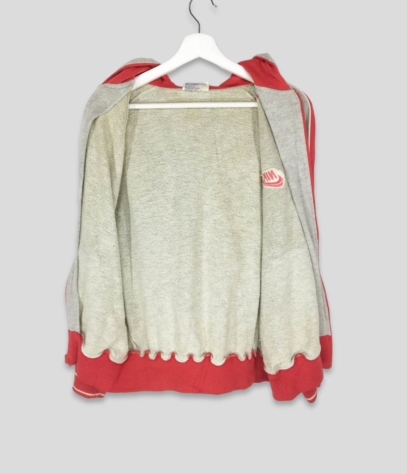 K&ouml;nnte beinhalten: Ein grauer Kapuzenpullover mit Rei&szlig;verschluss und roten Akzenten, der an einem wei&szlig;en Kleiderb&uuml;gel h&auml;ngt. Die Kapuze und der Saum sind rot, mit einem wei&szlig;en Streifen. Das Nike-Logo ist auf der Brust zu sehen. Die Jacke ist offen.