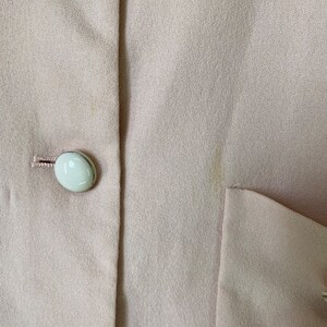 May include: A close-up of a light pink blazer with white buttons. The blazer has a smooth texture and a single pocket on the right side.