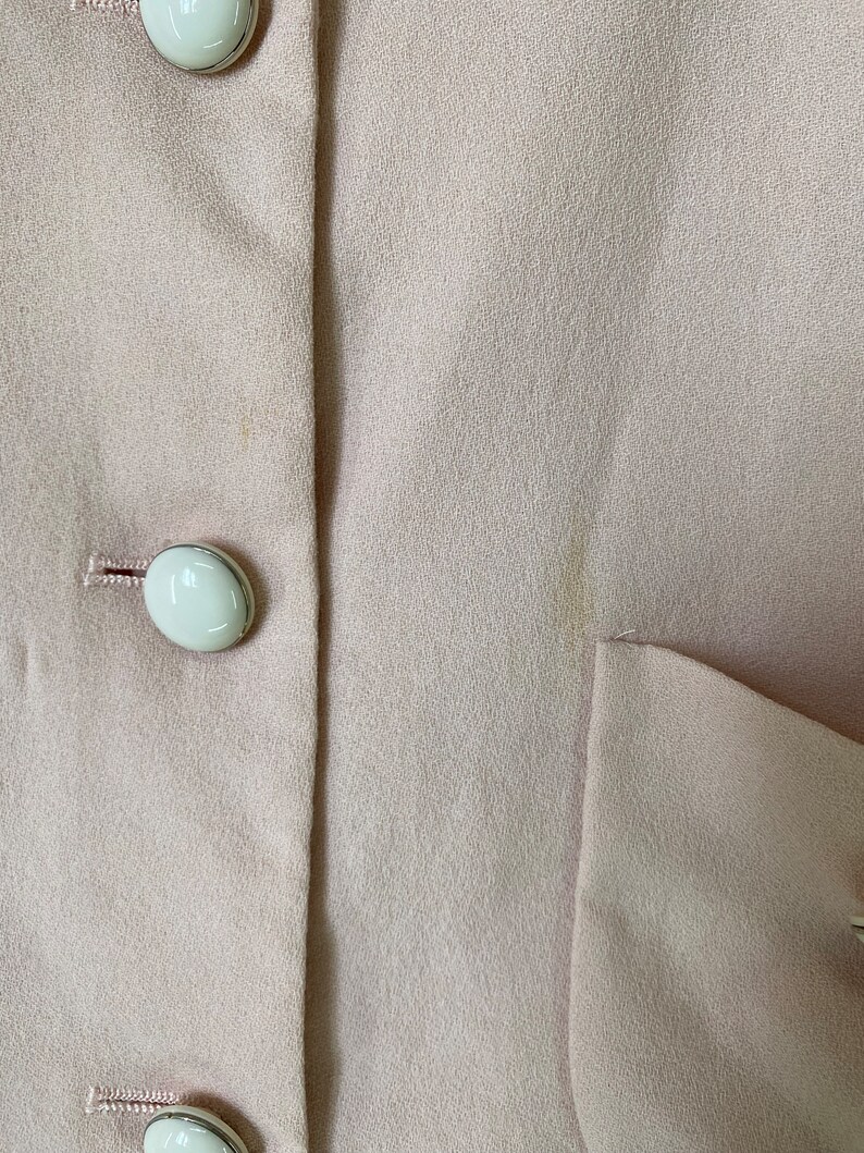 May include: A close-up of a light pink blazer with white buttons. The blazer has a smooth texture and a single pocket on the right side.