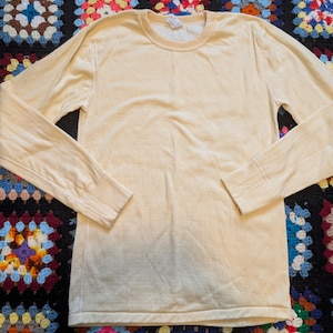 May include: A long-sleeved, cream-coloured shirt with a crew neck and ribbed cuffs. The shirt has a slightly faded appearance, suggesting a vintage or worn aesthetic. The fabric appears to be soft and comfortable.