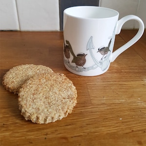 Peut inclure: Deux galettes rondes et brun clair sur une surface en bois à côté d'une tasse en céramique blanche avec une ancre bleue et des oiseaux bruns portant des chapeaux de marin.