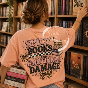 Pu&ograve; includere: Una maglietta color pesca con la scritta "SPICY BOOKS AND EMOTIONAL DAMAGE" in un font retr&ograve;. Il design include fiocchi con stampa leopardata e una pila di libri. La persona tiene in mano una pila di libri.