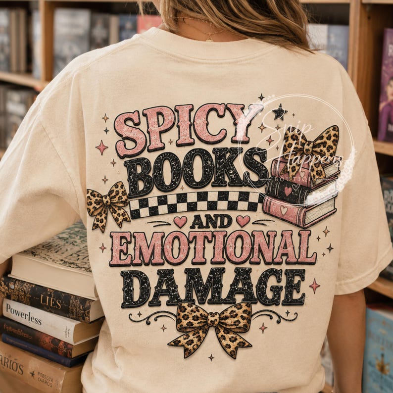 Pu&ograve; includere: T-shirt color crema con la scritta "Spicy Books and Emotional Damage" in rosa e nero. Il design include fiocchi con stampa leopardata, una pila di libri e un motivo a scacchi.