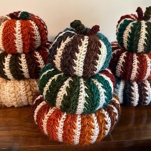 May include: Three stacks of crocheted pumpkins in various sizes, featuring vertical ribbed textures in autumn colors including orange, dark green, brown, cream, and burgundy. Each pumpkin is topped with a small, dark green crocheted stem.