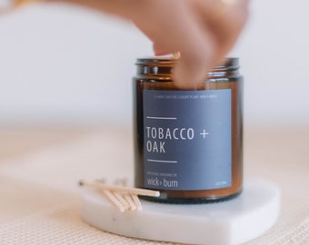 May include: A brown glass candle jar with a gray label that reads "Tobacco + Oak" and "wick + burn". The candle is on a white marble coaster.
