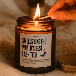 May include: A lit candle in a brown glass jar with a gold lid. The label reads "SMELLS LIKE THE WORLD'S BEST LASH TECH." A hand is holding a lit match over the candle. The candle is on a soft, textured surface.