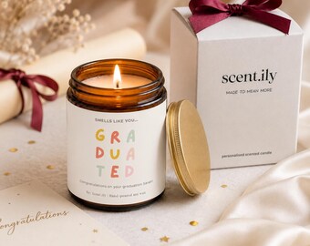 May include: A lit soy wax candle in an amber glass jar with a white label reading "SMELLS LIKE YOU... GRADUATED" and "Congratulations on your graduation Sarah!". Beside it sits a gold lid and a white gift box with a dark red ribbon bow on top.
