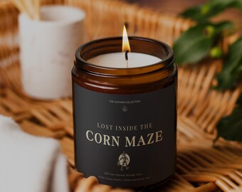 May include: A lit candle in a brown glass jar with a black label that reads "LOST INSIDE THE CORN MAZE". The candle is part of "THE AUTUMN COLLECTION" and sits on a woven basket. A small white ceramic container with matches is in the background.