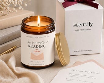 May include: A lit soy wax candle in an amber glass jar with a label reading Smells like you're going to... The University of READING, ACCEPTED!, and Well done Sophie xxx. Beside it sits a gold lid, a white gift box with a maroon ribbon, and a letter.
