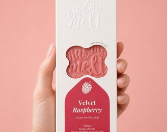 May include: A rectangular white cardboard box containing pink wax melts, labeled "make me melt" and "Velvet Raspberry." The packaging features a cutout window revealing the embossed wax, a raspberry graphic, and text stating "Hand-poured scented wax melts," "Made with soy wax," "Vegan," "Small batch," and "Hand-poured in England."