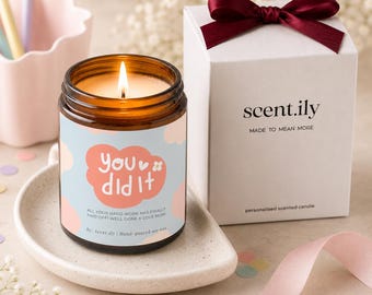 May include: A lit candle in an amber glass jar with a light blue and pink cloud-patterned label reading "you did it" and "all your hard work has finally paid off! well done x love mum!" next to a white gift box with a dark red ribbon bow.