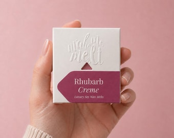May include: A square white cardboard box featuring the embossed text "make me melt" above a triangular cutout. A dark magenta label is applied to the lower portion, displaying the white text "Rhubarb Creme" and "Luxury Soy Wax Melts".