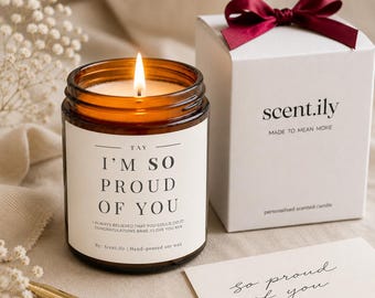 May include: A lit candle in an amber glass jar with a white label reading I'M SO PROUD OF YOU, next to a white gift box with a dark red ribbon and a card with the handwritten text so proud of you. The candle is hand-poured soy wax by Scent.ily.