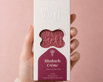 May include: A rectangular white cardboard box containing pink wax melts with the embossed text make me melt. The packaging features the text Rhubarb Crème, Luxury Soy Wax Melts, Vegan, Small Batch, and Hand-Poured in England on a dark pink base.