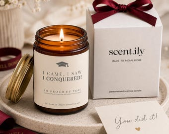 May include: A lit soy wax candle in an amber glass jar with a label reading I CAME, I SAW I CONQUERED! SO PROUD OF YOU! next to a white gift box with a burgundy ribbon, a gold lid, and a small card with the text You did it! and a heart icon.
