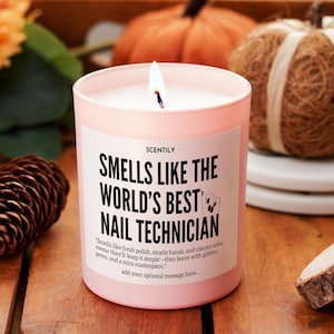Op de afbeelding: Een brandende roze kaars met de tekst "Smells Like The World's Best Nail Technician". De kaars zit in een roze pot met een wit etiket. De achtergrond bevat een dennenappel en decoratieve pompoenen.