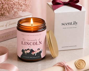 May include: A scented candle in an amber glass jar with a pink label featuring the text "SMELLS LIKE YOU'RE GOING TO... The University of LINCOLN" and an illustration of graduates. The candle has a gold lid and sits next to a white box with a maroon bow.