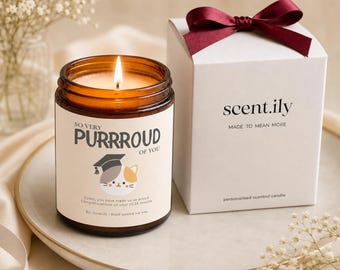 May include: An amber glass jar candle with a lit wick and a label reading SO VERY PURRROUD OF YOU with a cat wearing a graduation cap. Beside it sits a white gift box with a dark red ribbon, labeled scent.ily MADE TO MEAN MORE.