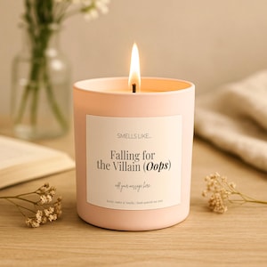 Pode incluir: Uma vela rosa claro num frasco de vidro cilíndrico com um pavio aceso. O rótulo branco apresenta o texto "SMELLS LIKE... Falling for the Villain (Oops) add your message here. Scent: Amber & Vanilla | Hand-poured soy wax" em preto.