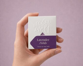 May include: A square white cardboard box featuring the embossed text "make me melt" at the top. A purple label is attached to the bottom half, displaying the white text "Lavender Fields" and "Luxury Soy Wax Melts" in a clean, sans-serif font.