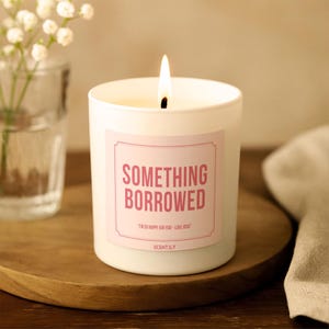 May include: A white cylindrical candle in a glass container with a lit wick. The label is light pink with a decorative border, featuring the text SOMETHING BORROWED in bold pink letters and the smaller text I'M SO HAPPY FOR YOU! - LOVE JOSIE.