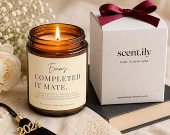 May include: A lit amber glass candle jar with a cream label reading Exams... COMPLETED IT MATE. BECCA AND COLE - WISHING YOU A LIFETIME OF LOVE, ENDLESS LAUGHTER, AND A PERMANENT BREAK FROM ESSAYS. Beside it sits a white gift box with a burgundy ribbon.