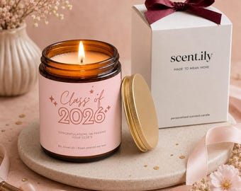 May include: A lit soy wax candle in an amber glass jar with a pink label reading Class of 2026, Congratulations on passing your GCSE's. Beside it sits a gold lid and a white gift box with a dark red ribbon, labeled scent.ily Made to Mean More.