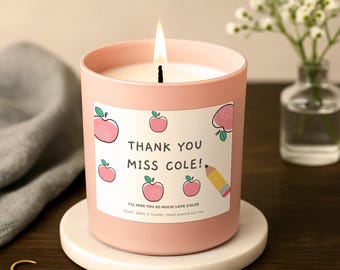Op de afbeelding: Een lichtroze kaars in een glazen pot met een wit etiket voorzien van roze appelillustraties, een potloodafbeelding en de tekst: THANK YOU MISS COLE! I'LL MISS YOU SO MUCH! LOVE CHLOE. Scent: Amber & Vanilla | Hand-poured soy wax.