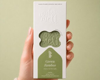 May include: A rectangular white box of hand-poured scented soy wax melts labeled Green Bamboo. The packaging features a cutout window revealing the sage green wax inside, which is embossed with the text make me melt in a cursive font.