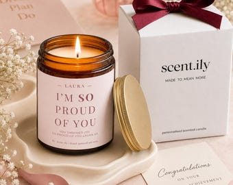 May include: A lit candle in an amber glass jar with a pink label reading I'M SO PROUD OF YOU, next to a white gift box with a dark red ribbon, a gold lid, and a card that reads Congratulations on your amazing achievement in black text.