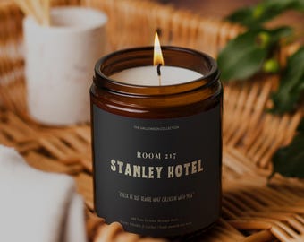 May include: A lit candle in a brown glass jar with a black label. The label reads "STANLEY HOTEL" and "ROOM 217" in gold lettering. The candle is part of "THE HALLOWEEN COLLECTION". The candle sits on a woven basket.