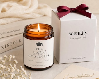 May include: A lit amber glass candle with a white label reading THE SWEET SMELL OF SUCCESS and a graduation cap icon, next to a white gift box with a dark red ribbon, a cream card with the word Congratulations, and a gold pen.