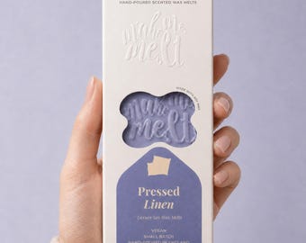 May include: A rectangular white cardboard box of hand-poured scented wax melts. The packaging features the text make me melt, a cutout window revealing light purple wax, and the label Pressed Linen with the text Vegan, Small Batch, Hand-Poured in England.