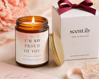 May include: A lit candle in an amber glass jar with a label reading AVA I'M SO PROUD OF YOU LOTS OF LOVE NAN AND GRANDAD XX. Beside it sits a gold metal lid and a white gift box with a dark red ribbon, labeled scent.ily MADE TO MEAN MORE.