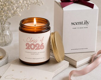 May include: A lit soy wax candle in an amber glass jar with a label reading Class of 2026 Congratulations on passing your GCSE's. Beside it sits a white gift box with a dark red ribbon, a gold metal lid, and a card with the word Congratulations.